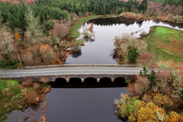 Naklejka premium Aerial view of river winding through autumnal forest. Bridge crosses calm water over grassy banks. Trees show fall colors: red, orange, brown. Green patches contrast with seasonal hues.