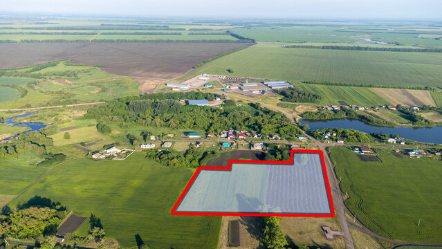 Aerial view captures a rural farm landscape. Lush green fields stretch across the horizon. Red outline marks a specific plot of land. Nearby structures and water bodies frame the scene
