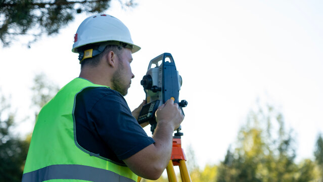 Man in white hard hat and green safety vest uses theodolite. He concentrates on measuring land features. Bright sky and trees form natural outdoor backdrop