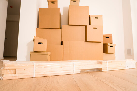 Stacked cardboard boxes fill a room. Various-sized boxes create a storage display. Wooden pallet rests on light wood flooring. White walls and a hallway entrance complete the setting