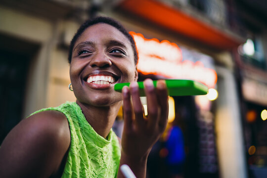 Close up of smiling African American woman holding mobile phone near her face for voice chat. Joyful black female student reading notifications or looking away with wide radiant smile.