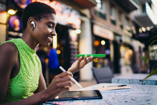 Cheerful African American woman recording voice message or chatting on speakerphone near cafe terrace table. Happy black female student using green smartphone for online communication in city.
