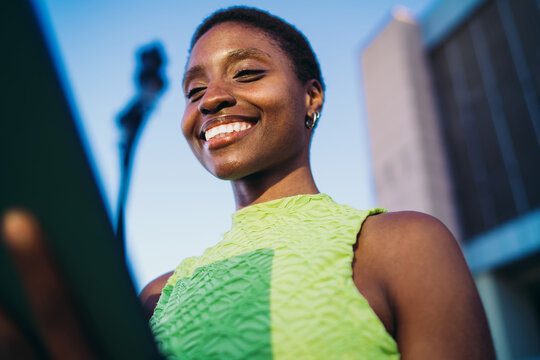 Low angle portrait of happy African American woman laughing with tablet device at sunset. Cheerful dark skinned model checking online money transfers or scrolling media feed.