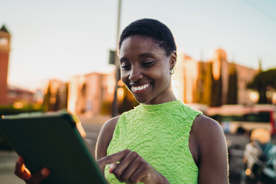 Cheerful dark skinned woman tapping on tablet touch screen against golden hour sunset background. Happy African American female traveler scrolling social media or sending money online.