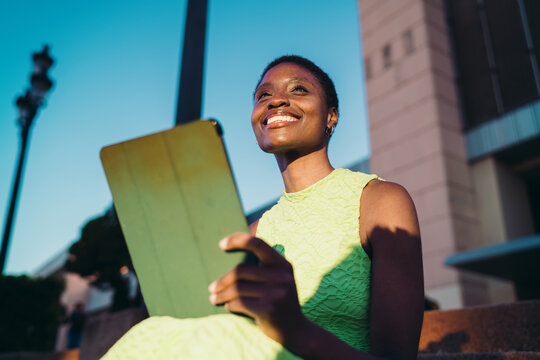Happy dark skinned woman in neon dress holding tablet and looking away with radiant smile. Cheerful African American youth receiving positive notification or social media chat alert.