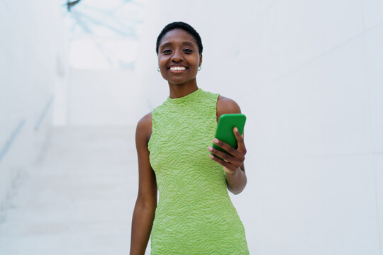 portrait of cheerful African American woman in green dress, looking directly at the camera with radiant smile. She holds smartphone and tablet against clean white minimalist background