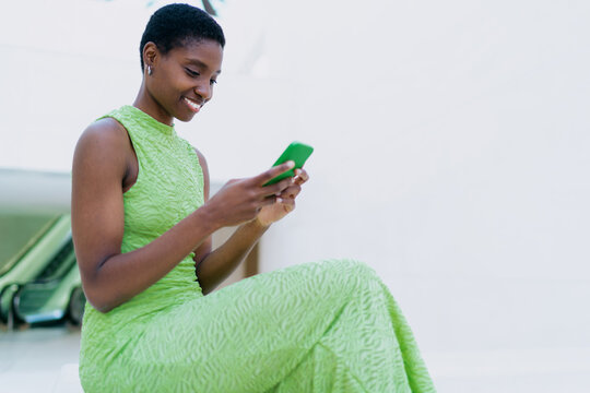 Smiling African American woman holding a green smartphone and a digital tablet against a minimalist white background. Dark skinned model wearing a light green open back dress, looks away.