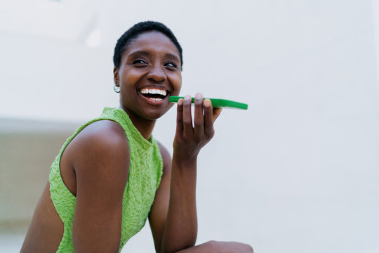 Joyful African American woman text messaging on mobile phone. Cheerful black female youth chatting on smartphone screen app, looking away with toothy smile near minimalist backdrop.