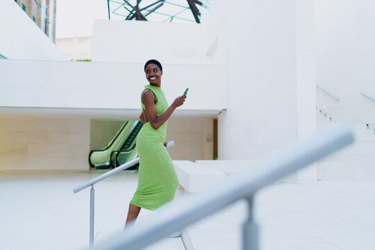 Happy African American woman looking back with smile on modern stairs. Cheerful dark skinned female traveler holding smartphone, checking transport schedule or digital booking notifications.