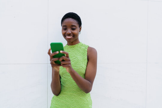African American woman using mobile application, typing message. Cheerful Black female scrolling social media network on smartphone screen near white wall with copy space, doing freelance remote work.