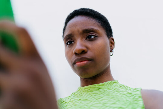 Close up portrait of beautiful black female model looking down at screen, holding smartphone near face. Focused African American woman student reading text chat message near white background wall
