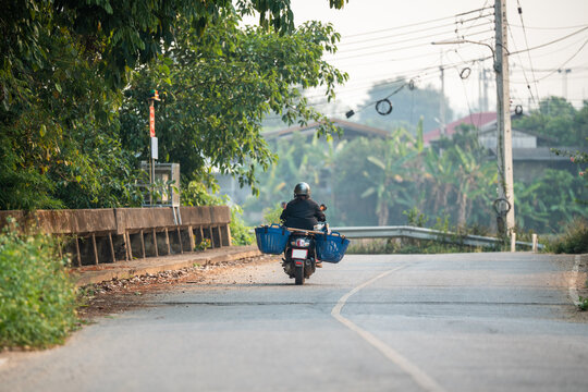 Unrecognizable local Thai farmer in helmet driving on motorcycle carrying plastic baskets with harvest, man worker riding along countryside road on scooter with containers of produce in Thailand.