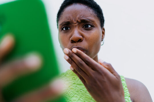 Extreme close up view of surprised dark skinned model looking down at smartphone screen. Pensive African American woman face reading unexpected chat notification near white wall negative space