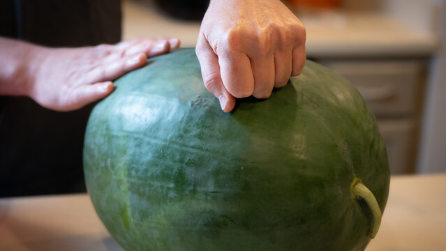 person knocking on a whole, round green black diamond watermelon on a countertop