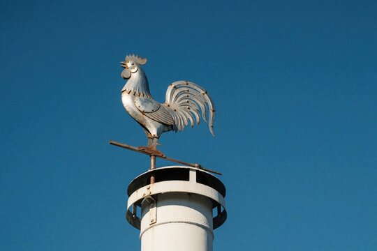 Metal rooster weather vane sits on white chimney pipe. Old iron bird decoration stands against clear blue sky. Rustic farmhouse roof accessory shows vintage style architecture. Traditional wind vane