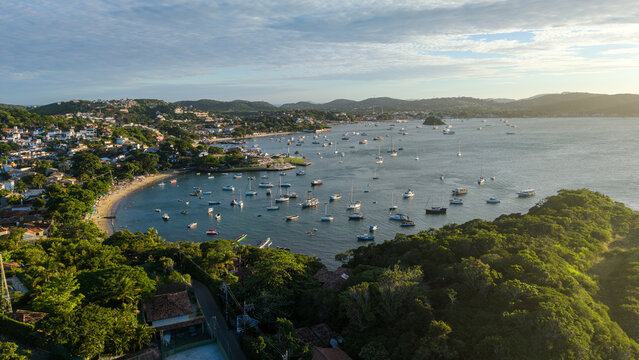 Aerial view of Praia dos Ossos with numerous boats anchored in the bay, surrounded by lush green hills and coastal architecture in Armacao dos Buzios, Rio de Janeiro, Brazil.