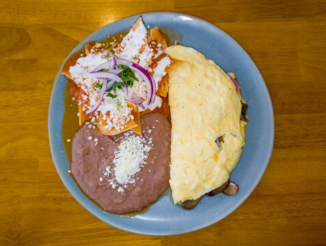 A plate featuring a folded omelet filled with mushrooms next to a portion of chilaquiles topped with red sauce, crema, cheese, and red onion slices, alongside refried beans sprinkled with cheese.