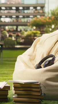 Portrait format: Stacked books and headphones sit on a soft beanbag in a sunny outdoor campus space, creating empty academic study setup for education and learning content, reflecting student life.