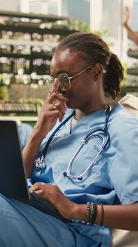 Vertical aspect: The portrait shows a woman student smiling and typing on a laptop from a beanbag in a modern outdoor campus courtyard, with a med school peer nearby in blue scrubs and a stethoscope.