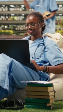 Vertical aspect: The portrait shows a woman student smiling and typing on a laptop from a beanbag in a modern campus courtyard during outdoor medical study.