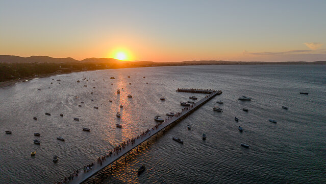 Aerial view of Manguinhos pier extending into the bay with numerous small boats anchored in the calm water during a golden sunset in Armacao dos Buzios, Rio de Janeiro, Brazil.