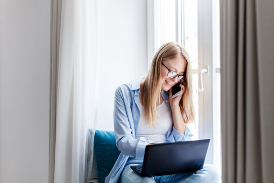 Young woman working from home office. Freelancer using laptop and talking on mobile phone. Workplace in living room on windowsill. Technology lifestyle. Concept of female business, teleworking