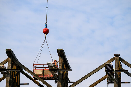 Workman in safety equipment in work basket help aloft by crane, old wood railway train trestle construction project to update structural framework, sunny summer transportation project, United States

