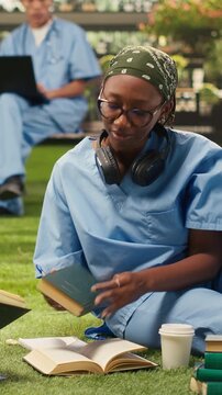 Vertical screen: Women medical students in blue scrubs compare open textbooks, discussing coursework on a modern campus lawn, group study notes and academic materials nearby.