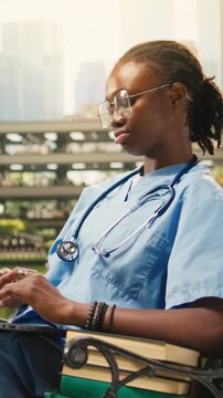 Vertical aspect: The woman medical student types on a laptop while seated on a bench on a modern campus courtyard for outdoor learning, with the young adult studying in blue scrubs and a stethoscope.
