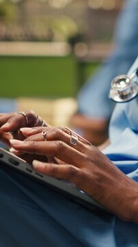 Vertical frame: Close-up: a medical student typing on a laptop keyboard from a beanbag in bright modern campus courtyard outdoors, with the woman wearing scrubs and a stethoscope for medical school.