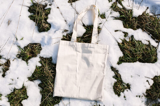 White cotton mesh bag on spring snow, green grass thawed patches background. Zero waste, no plastic, eco friendly shopping, recycling concept. Blank mockup shopper, place for text. copy space flat lay