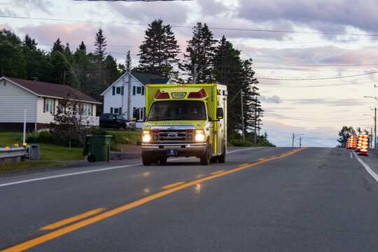 Gaspesie, Quebec, 15 July 2026 : A bright yellow Ford ambulance drives on a paved road with its headlights on, passing residential houses and trees.