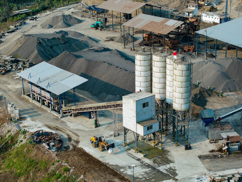 Aerial view of a concrete batching plant with tall storage silos, piles of gravel and sand, and a front loader operating at an industrial site in INDONESIA.