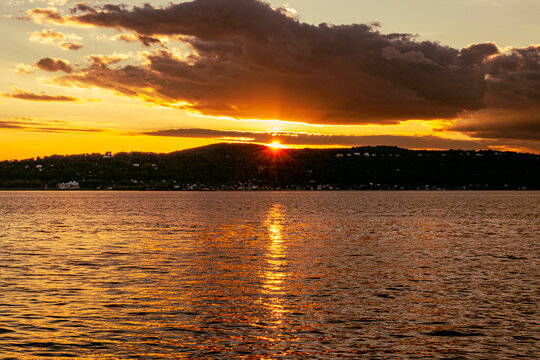 A golden sunset over the Hudson River casts warm light across rippling water, with the sun glowing above a dark wooded shoreline and dramatic clouds filling the evening sky.