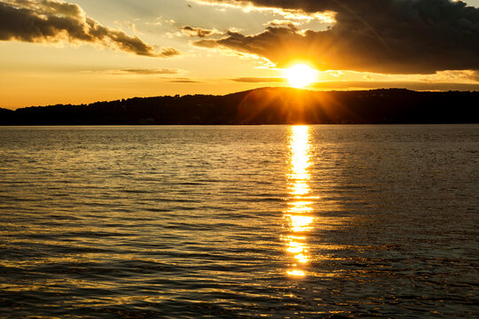 A golden sunset over the Hudson River casts warm light across rippling water, with the sun glowing above a dark wooded shoreline and dramatic clouds filling the evening sky.