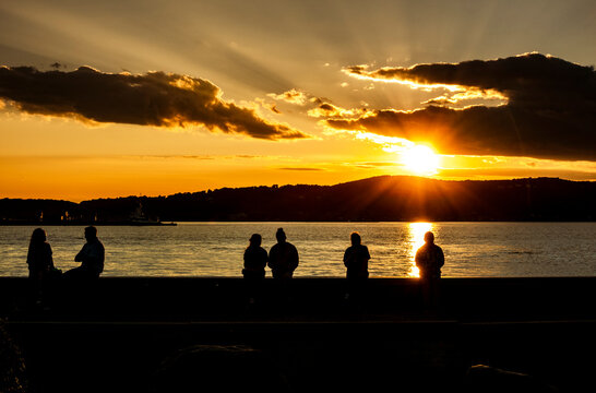 A breathtaking sunset over the Hudson River casts golden light across the water, while silhouetted hills and people along the shoreline create a peaceful, dramatic evening scene beneath golden clouds.