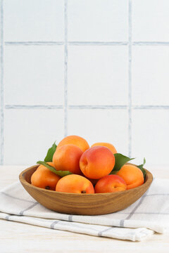 Fresh ripe apricots arranged in a wooden bowl on white kitchen table and tiled wall background with copy space. Summer stone fruits with green leaves, rustic home kitchen setting, healthy food