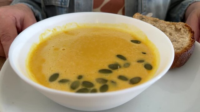White bowl of creamy pumpkin soup with green seeds and slice of bread on plate, person at table. Healthy lunch, vegetarian meal, autumn dish