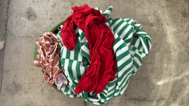 Green basin with pile of colorful laundry and striped towel on concrete floor. Wash day, household chores, dirty clothes, home washing