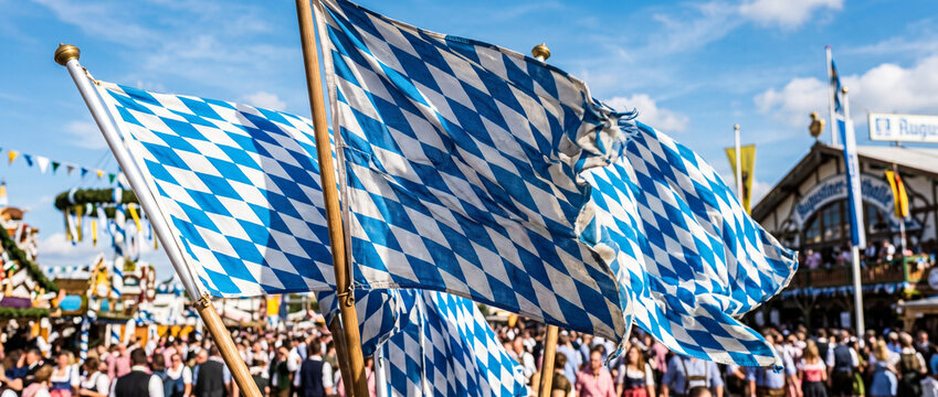 Bavarian flags waving above a crowded Oktoberfest fairground in Munich. Traditional German festival celebration with colorful decoration, outdoor beer culture, and cheerful tourism atmosphere.