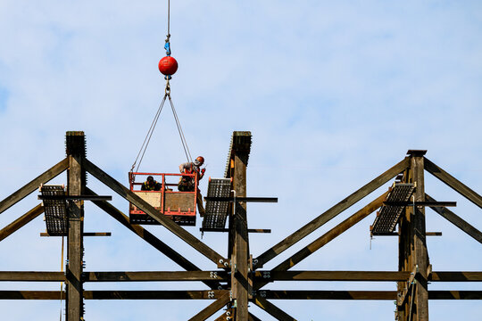 Construction worker moving from crane work basket to scaffolding on old wood train trestle, structural framework rebuild, handheld radio communication, sunny summer day, America
