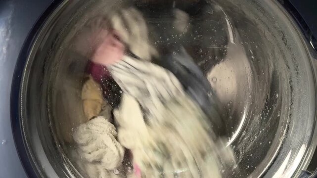 Colorful laundry spinning inside washing machine drum behind wet glass door. Wash day, household chores, cleaning clothes, home appliance in use