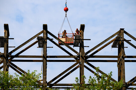 Worker in safety equipment in work on scaffolding overhead, old wood railway train trestle construction project to update structural framework, sunny summer transportation project, United States

