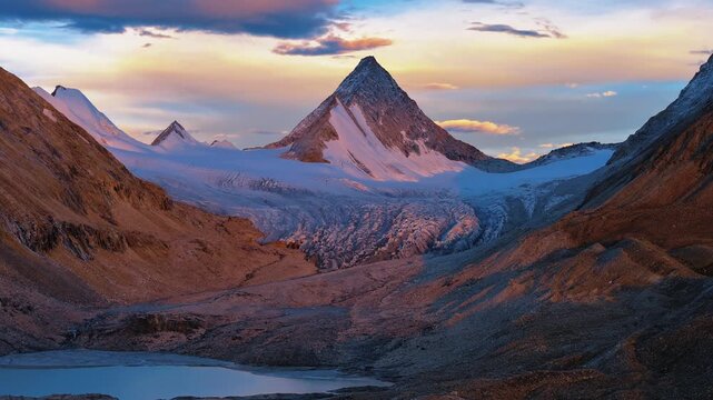 Cinematic view of dramatic snow-covered mountain peaks surrounded by a vast glacier valley and rugged alpine landscape. Warm sunlight illuminates the mountains beneath a changing colorful sky, creatin