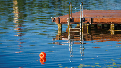 Wooden swimming pier with a metal ladder and an orange buoy on a calm lake with blue water on a sunny day. © Dmytro