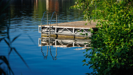 Floating wooden pier with a metal ladder on a calm blue lake, framed by green bushes and leaves on a sunny day. © Dmytro