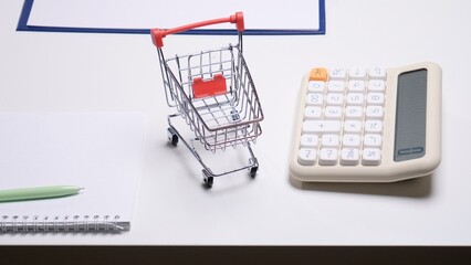 Miniature shopping cart placed next to a calculator and spiral notebook on a white desk, symbolizing retail finance management, consumer spending, and budget tracking © Aliaksandr