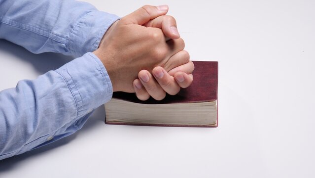 Person's hands clasped in prayer resting on a traditional closed book, symbolizing faith, devotion, spirituality, and seeking solace or guidance from religious texts