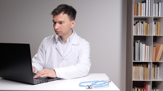 Medical professional typing on a laptop, conducting research or telehealth consultation, with a stethoscope resting on the white desk beside a bookshelf