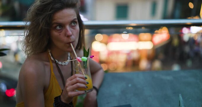 A young woman in a yellow top with a seashell necklace sips a refreshing cocktail from a glass with a straw, enjoying the evening ambience with blurred city lights in the background.
